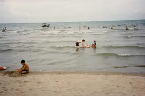 Đây là một khung cảnh tại một bãi biển vào một ngày nhiều mây. Rất đông người, bao gồm cả người lớn và trẻ em, đang tắm biển và vui chơi. Ở phía trước, trên bãi cát, một cậu bé đang ngồi chơi với cát, có vẻ như đang xây lâu đài cát. Cạnh cậu bé có một chiếc phao cứu sinh màu đỏ và trắng. Dưới nước, ở khu vực gần bờ, một người đàn ông đang chơi cùng hai đứa trẻ nhỏ với một chiếc phao cứu sinh khác. Xa hơn ngoài biển, nhiều người khác cũng đang bơi lội. Có thể thấy một chiếc thuyền gỗ nhỏ có mái che đang neo đậu ở phía xa. Không có văn bản nào có thể đọc được trong hình ảnh.。（日本語訳）。これは曇りの日のビーチの光景です。大人も子供も含むたくさんの人が、海水浴をしたり遊んだりしています。手前の砂浜では、一人の少年が砂で遊んでおり、砂の城を建てているようです。少年の隣には、赤と白の浮き輪があります。水の中、岸に近いエリアでは、一人の男性が別の浮き輪を使って二人の小さな子供と遊んでいます。さらに沖では、他の多くの人々も泳いでいます。遠くに、屋根付きの小さな木造船が停泊しているのが見えます。画像内に読み取れるテキストはありません。