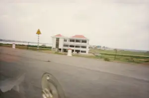 Bức ảnh chụp lại khung cảnh ven một con đường nhựa, có lẽ được chụp từ bên trong một chiếc ô tô đang di chuyển. Bên lề đường có một biển báo giao thông nguy hiểm hình tam giác màu vàng trên cột sọc trắng đen, nhưng không thể nhìn rõ ký hiệu bên trong. Gần đó là các cột mốc bê tông sơn trắng với phần đỉnh màu đỏ. Nổi bật trong ảnh là một công trình xây dựng lớn, một tòa nhà nhiều tầng sơn trắng, lợp mái ngói đỏ, có vẻ như đang trong giai đoạn hoàn thiện vì các ô cửa sổ còn để trống. Xung quanh tòa nhà là không gian thoáng đãng với những cánh đồng xanh ở phía bên phải. Ở phía xa bên trái là một vùng nước rộng lớn, có thể là sông, hồ hoặc bờ biển. Góc dưới bên trái của ảnh có một phần của chiếc xe, giống như nắp chụp mâm bánh xe, bị mờ do chuyển động.。（日本語訳）。写真はアスファルト道路沿いの風景を撮影したもので、おそらく移動中の車内から撮られたものでしょう。路肩には、白黒の縞模様のポールに黄色い三角形の危険を示す交通標識がありますが、中の記号ははっきりと見えません。その近くには、上部が赤く塗られた白いコンクリートのキロポストがあります。写真の中で目立っているのは大きな建設中の建物で、白い塗装の多層階建て、赤い瓦屋根で、窓枠が空いていることから、完成間近の段階にあるようです。建物の周りは広々とした空間で、右側には緑の田畑が広がっています。遠く左側には、川、湖、あるいは海岸かもしれない広大な水域があります。写真の左下隅には、ホイールキャップのような車の一部が写っており、動きによってぼやけています。