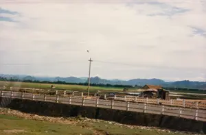 Bức ảnh chụp một khung cảnh nông thôn ở Việt Nam vào khoảng năm 1997. Ở tiền cảnh là một con đường được xây trên cao, có lan can bê tông màu trắng với các vạch sơn đỏ và trắng xen kẽ. Trên đường, có một biển báo màu xanh lam với chữ trắng nhưng không thể đọc rõ nội dung. Có một vài người đang di chuyển trên đường và một nhóm người tụ tập tại một ngôi nhà lợp ngói đơn sơ bên phải, có thể là một quán ven đường. Phía sau con đường là một vùng đất nông nghiệp rộng lớn, bao gồm các cánh đồng xanh và các thửa ruộng ngập nước, có thể là ruộng lúa hoặc ruộng muối. Giữa cánh đồng có một túp lều nhỏ. Ở phía xa là dãy núi trập trùng ẩn hiện trong mây. Một cột điện và đường dây điện chạy ngang qua bức ảnh, đặc trưng cho cảnh quan nông thôn thời kỳ này.。（日本語訳）。この写真は1997年頃のベトナムの田舎の風景を写したものです。前景には、赤と白の縞模様に塗装された白いコンクリートの欄干がある、高架の道路があります。道路上には、白い文字が書かれた青い看板がありますが、内容ははっきりと読み取れません。道路上には数人が移動しており、右側には簡素な瓦葺きの家に集まっている人々のグループがいます。これは道端の店かもしれません。道路の後ろには広大な農地が広がっており、緑の畑や水田、あるいは塩田と思われる水が張られた田んぼが含まれています。畑の真ん中には小さな小屋があります。遠くには、雲間に見え隠れする山々が連なっています。電柱と電線が写真を横切っており、この時代の田舎の風景を特徴づけています。