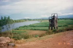 Trên ảnh có dấu ngày tháng màu cam: 97 7 14.。Bức ảnh ghi lại một khung cảnh nông thôn ở Việt Nam. Cảnh vật bao gồm một con sông chảy ở phía bên trái, bao quanh bởi những cánh đồng nông nghiệp xanh tươi, có cả những thửa ruộng đất đã được cày xới. Ở phía trước là một con đường đất.。Điểm nổi bật trong ảnh là một công trình bê tông bị bỏ hoang và hư hỏng nặng, có thể là một lô cốt hoặc tháp canh còn sót lại từ thời chiến. Công trình này đứng trên các cột trụ và có nhiều lỗ thủng trên tường.。Phía xa là những rặng cây và dãy núi mờ ảo dưới bầu trời nhiều mây. Không có người hay hoạt động nào được ghi lại, chỉ có một phong cảnh tĩnh lặng.。（日本語訳）。写真にはオレンジ色の「97 7 14」という日付スタンプがあります。この写真はベトナムのある田園風景を記録したものです。風景には、左側を流れる川、その周りに広がる青々とした農地、そして耕された畑が含まれています。手前には未舗装の道があります。写真の中で際立っているのは、放置されひどく損傷したコンクリートの建造物で、これは戦時中から残っているトーチカか監視塔かもしれません。この建造物は柱の上に立っており、壁には多くの穴が開いています。遠くには、曇り空の下に木々の列とぼんやりとした山脈が見えます。人や活動は記録されておらず、ただ静かな風景が広がっています。