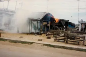 Một vụ hỏa hoạn đang xảy ra tại một khu nhà tạm hoặc gian hàng ven đường. Một trong những công trình, có vẻ như có khung bằng gỗ và mái lợp tôn, đang bốc cháy. Ngọn lửa màu cam sáng đang bùng lên từ phần mái, trong khi khói trắng dày đặc bốc ra từ phía bên trái của tòa nhà. Phía trước, trên nền đất, có những chiếc bàn và ghế gỗ trống rỗng, gợi ý đây có thể là một khu chợ hoặc khu vực kinh doanh nhỏ. Một đống cát nhỏ và vài viên gạch cũng nằm rải rác. Ở phía sau, có thể nhìn thấy một chiếc xe tải màu xanh có bạt che, các tòa nhà khác và hệ thống dây điện chằng chịt trên đầu. Không có chữ viết nào có thể đọc được. Toàn bộ khung cảnh cho thấy một sự kiện phá hủy đang diễn ra.。（日本語訳）。路上にある仮設住宅または屋台で火災が発生しています。その建物の一つは、木製の骨組みとトタン屋根でできているように見え、燃えています。明るいオレンジ色の炎が屋根の部分から燃え上がっており、一方で建物の左側からは濃い白煙が立ち上っています。手前の地面には、誰も座っていない木製のテーブルと椅子があり、ここが市場か小規模な商業エリアであった可能性を示唆しています。小さな砂の山といくつかのレンガも散らばっています。後方には、幌で覆われた青いトラック、他の建物、そして頭上には複雑に絡み合った電線が見えます。読み取れる文字はありません。全体の光景は、破壊的な出来事が進行中であることを示しています。