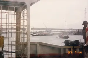 Chữ viết duy nhất có thể đọc được là dấu ngày tháng "97 7 14".。Bối cảnh là một khu vực sông nước, có thể là một bến cảng hoặc bến phà vào một ngày nhiều mây hoặc có mưa. Bức ảnh được chụp từ trên một con tàu hoặc phà lớn, với góc nhìn qua một khung lưới sắt ở bên trái và một phần lan can bê tông. Ở phía bên phải, một người đàn ông đội mũ cối đang đứng.。Trên mặt sông, có nhiều ghe, tàu đang hoạt động. Nổi bật là một chiếc ghe lớn có mui che, trên đó có một người đội nón lá đang đứng. Một chiếc ghe nhỏ hơn cũng đang di chuyển gần đó.。Ở hậu cảnh, một cây cầu bê tông lớn bắc qua sông. Gần cây cầu có thể thấy các cần cẩu, cho thấy đây có thể là một khu vực cảng hoặc công trường xây dựng. Nhiều tàu thuyền khác đang neo đậu dọc bờ. Ngoài ra còn có các cột đèn đường và một cột tháp lưới thép ở phía xa.。（日本語訳）。読み取れる唯一の文字は「97 7 14」という日付の印です。背景は水辺の地域で、曇りか雨の日の港またはフェリー乗り場である可能性があります。写真は大きな船またはフェリーの上から撮影されたもので、左側には鉄格子のフレーム、そしてコンクリートの手すりの一部を通して見た視点です。右側には、ヘルメットをかぶった男性が立っています。川面では、多くの小舟や船が行き交っています。目立っているのは屋根付きの大きな小舟で、その上にはノンラー（菅笠）をかぶった人が立っています。その近くでは、より小さな小舟も動いています。背景には、大きなコンクリートの橋が川にかかっています。橋の近くにはクレーンが見え、ここが港湾地区または建設現場である可能性を示しています。他の多くの船が岸に沿って停泊しています。さらに、遠くには街灯の柱と鉄骨の塔もあります。