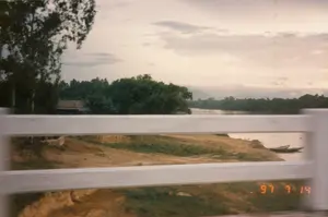 Bức ảnh chụp phong cảnh một vùng quê ven sông, có khả năng được chụp từ một phương tiện đang di chuyển qua cầu. Khung cảnh chính là một con sông rộng, nước chảy êm đềm. Ven bờ sông là một bãi đất với cỏ mọc thưa thớt, nơi có hai chiếc thuyền gỗ nhỏ (ghe) đang neo đậu sát mép nước. Phía bên kia sông là một dải cây cối xanh um tùm, che khuất một phần ngôi nhà có mái ngói. Xa xa phía chân trời là hình ảnh mờ của những dãy núi. Bầu trời u ám, có nhiều mây. Phần tiền cảnh của bức ảnh bị che bởi các thanh ngang màu trắng, dường như là lan can của cây cầu. Ở góc dưới bên phải có dấu ngày tháng kỹ thuật số màu cam ghi '97 7 14'.。（日本語訳）。川沿いの田舎の風景を撮影した写真で、橋を渡っている乗り物から撮影された可能性がある。主な風景は、穏やかに流れる広い川である。川岸には草がまばらに生えた土地があり、そこには2隻の小さな木造船（ghe）が水際に係留されている。川の向こう側には緑豊かな木々が連なり、瓦屋根の家の一部を覆い隠している。空はどんよりとしており、雲が多い。写真の前景は、橋の欄干と思われる白い横棒によって遮られている。右下隅には、「'97 7 14」と記されたオレンジ色のデジタル日付スタンプがある。