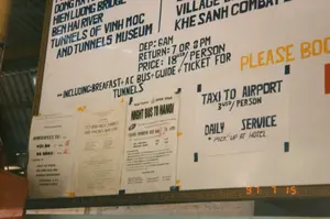 右下のデジタル日付スタンプは、写真が1997年7月15日に撮影されたことを示しています。掲示板には、英語で手書きおよびタイプされた多くの情報が含まれており、観光ツアーや交通サービスを宣伝しています。掲示板のテキスト内容には以下が含まれます：。1.  歴史的な場所を巡るツアーを宣伝する大きな看板：「ドンハ、ヒエンルオン橋、ベンハイ川、ヴィンモックトンネルとトンネル博物館、ケサン戦闘基地」。*   スケジュール：「出発：午前6時、帰着：午後7時または8時」（出発：午前6時、帰着：午後7時または8時）。*   料金：「価格：18米ドル/人」（料金：1人あたり18米ドル）。*   含まれるもの：「朝食＋エアコンバス＋ガイド＋トンネル入場券を含む」（含まれるもの：朝食＋エアコンバス＋ガイド＋トンネル入場券）。2.  「空港へのタクシー」（空港へのタクシー）サービスに関する手書きの告知で、料金は「3米ドル/人」（1人あたり3米ドル）、毎日運行（「DAILY SERVICE」）、ホテルでのピックアップ（「PICK UP AT HOTEL」）です。3.  「ミニバス行き先：」（ミニバス行き：）ホイアンへは5米ドル、ダナンへは4米ドルで、「ランコービーチ、ハイヴァン峠、マーブルマウンテン」（ランコービーチ、ハイヴァン峠、五行山）に立ち寄るという広告用紙。4.  ハノイ・トセルコ社の「ハノイ行き夜行バス」（ハノイ行き夜行バス）の広告で、「ハノイ - ニンビン - フエ（毎日）」路線が22米ドル、午後6時出発。5.  「ヴィンモックトンネルとフォンニャ洞窟へ」（ヴィンモックトンネルとフォンニャ洞窟へ）のツアーに関する別の告知。全体の光景は、1990年代のベトナムの主要都市で一般的だった、観光客、特に外国人観光客にサービスを提供する場所を示しています