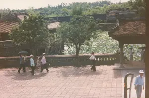 Bức ảnh chụp một khung cảnh tại một khu di tích lịch sử, có khả năng là ở Huế, vào khoảng năm 1997. Bối cảnh chính là một khoảng sân rộng lát gạch, nơi có nhiều người đang đi dạo tham quan.。Ở hậu cảnh là một hồ sen lớn với nhiều lá và hoa sen đang nở rộ. Bao quanh hồ là các công trình kiến trúc cổ của Việt Nam như nhà rường, đình nghỉ chân với mái ngói âm dương màu đỏ và các bức tường cũ. Có nhiều cây xanh mọc xen kẽ, và phía xa là một ngọn đồi rậm rạp cây cối.。Về hoạt động, có nhiều người, dường như là du khách, đang đi bộ trong sân. Một nhóm người bên trái gồm một người đang che ô đen, một phụ nữ mặc áo hồng đội mũ đang dắt tay một đứa trẻ, và những người khác. Một người phụ nữ mặc áo trắng và quần tối màu đang đi một mình ở khu vực giữa. Dưới một mái đình nghỉ chân ở bên phải, có một vài người đang đứng. Một người đàn ông đội mũ lưỡi trai trắng đang đi ở góc dưới bên phải. Trang phục của mọi người cho thấy đây là một ngày nắng nóng.。（日本語訳）。この写真は、1997年頃、フエと思われる歴史的遺跡の一場面を撮影したものです。主な背景は、多くの人々が散策し観光している、レンガ敷きの広い中庭です。後景には、たくさんの葉と満開の蓮の花がある大きな蓮池があります。池の周りには、伝統的な木造家屋（ニャールオン）、赤い陰陽瓦の屋根を持つ休憩用の東屋、そして古い壁など、ベトナムの古い建築物があります。多くの緑の木々が点在しており、遠くには木々が生い茂る丘があります。人々の活動については、観光客と思われる多くの人々が中庭を歩いています。左側のグループには、黒い傘をさしている人、ピンクの服を着て帽子をかぶり子供の手を引いている女性、そして他の人々がいます。中央エリアでは、白いシャツと暗い色のズボンを履いた女性が一人で歩いています。右側の休憩用の東屋の下には、数人が立っています。右下隅では、白い野球帽をかぶった男性が歩いています。人々の服装は、この日が暑い晴れた日であることを示しています。