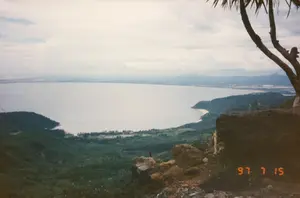 Trên hình có dấu ngày tháng màu cam '97 7 15'. Bức ảnh chụp một khung cảnh thiên nhiên hùng vĩ từ một điểm nhìn trên cao, rất có thể là từ Đèo Hải Vân. Phía dưới là một vùng vịnh hoặc đầm phá rộng lớn, mặt nước phẳng lặng. Bao quanh vịnh là những ngọn núi và sườn đồi được bao phủ bởi rừng cây xanh rậm rạp. Một dải đất ven biển hẹp chạy dọc theo bờ vịnh, có thể thấy lác đác cây cối và các công trình nhỏ. Ở phía xa, những dãy núi khác hiện ra mờ ảo trong sương. Bầu trời có nhiều mây. Ở tiền cảnh bên phải là một tảng đá lớn và bóng của một cành cây. Không có hoạt động nào của con người được ghi lại rõ ràng, chỉ có cảnh quan thiên nhiên yên bình.。（日本語訳）。写真にはオレンジ色の'97 7 15'という日付スタンプがあります。この写真は、高い視点から撮影された雄大な自然の風景で、おそらくハイヴァン峠からのものです。下には広大な湾またはラグーンがあり、水面は穏やかです。湾を囲んでいるのは、緑豊かな鬱蒼とした森に覆われた山々や丘陵です。湾岸に沿って細長い沿岸の土地が伸びており、まばらな木々や小さな建造物が見られます。遠くには、他の山脈が霧の中にぼんやりと現れています。空は曇っています。右の前景には、大きな岩と木の枝の影があります。人間の活動は明確に記録されておらず、ただ穏やかな自然の景観があるだけです。