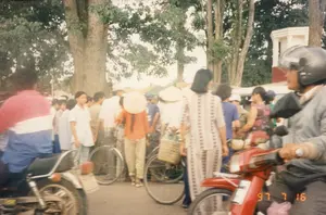 Bối cảnh là một con đường đông đúc người qua lại tại một khu vực có thể là thị trấn hoặc thành phố ở Việt Nam, được chụp vào ngày 16 tháng 7 năm 1997, như được chỉ ra bởi dấu ngày tháng '97 7 16' ở góc dưới bên phải bức ảnh. Một đám đông người dân đang tụ tập trên đường. Trong đám đông có nhiều người đi xe đạp và một số người đi xe máy. Có thể thấy một vài người phụ nữ đội nón lá, một người trong số họ mặc áo màu cam và đứng cạnh xe đạp. Một người phụ nữ khác mặc bộ đồ bộ (quần áo mặc ở nhà) có hoa văn. Một người mang theo chiếc giỏ đan bằng tre/mây, cho thấy hoạt động có thể liên quan đến việc đi chợ. Ở tiền cảnh, có hai chiếc xe máy: một chiếc màu đỏ bên phải do một người đàn ông đội mũ bảo hiểm điều khiển, và một chiếc khác ở bên trái. Dọc hai bên đường là những hàng cây cổ thụ lớn. Ở phía sau, có thể nhìn thấy một phần của một công trình kiến trúc màu trắng.。（日本語訳）。背景は、ベトナムの町か都市と思われる地域にある、人通りの多い道です。写真の右下隅にある「'97 7 16」という日付スタンプが示すように、1997年7月16日に撮影されました。大勢の人々が道に集まっています。人混みの中には、多くの自転車に乗る人々と、何人かのバイクに乗る人々がいます。ノンラー（菅笠）をかぶった女性が数人見え、そのうちの一人はオレンジ色の服を着て、自転車の隣に立っています。別の女性は、柄物の「đồ bộ」（部屋着）を着ています。ある人は竹か籐で編んだかごを持っており、市場へ行くことに関連する活動の可能性を示しています。前景には2台のバイクがあります。右側にはヘルメットをかぶった男性が運転する赤いバイクが1台、そして左側にもう1台あります。道の両脇には、大きな古い木々が並んでいます。後方には、白い建築物の一部が見えます。