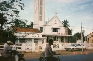 Bức ảnh chụp một nhà thờ Tin Lành ven đường vào ngày 16 tháng 7 năm 1997. Nhà thờ có kiến trúc hiện đại, sơn màu trắng với một tháp chuông cao và một cây thánh giá trên đỉnh mái. Trên mặt tiền có dòng chữ lớn màu đỏ ghi 'TIN LÀNH'. Phía trên cao hơn, trên phần mái tam giác, có biểu tượng một cuốn sách đang mở (Kinh Thánh) với hình quả địa cầu. Có thể đọc được chữ 'KINH THÁNH' trên biểu tượng này. Một tấm biển nhỏ hơn trên hàng rào có ghi chữ nhưng một phần bị mờ, chỉ đọc được 'CHI HỘI TIN LÁNH PHAN...'. Bao quanh nhà thờ là hàng rào bê tông. Bên cạnh là một tòa nhà phụ lợp mái ngói đỏ. Bên ngoài, một chiếc ô tô kiểu station wagon màu trắng đang đỗ. Trên con đường nhựa, người dân đang di chuyển bằng xe đạp, phản ánh cuộc sống thường ngày thời kỳ đó. Một người đàn ông đội mũ lưỡi trai đang đạp xe ở tiền cảnh.。（日本語訳）。1997年7月16日に撮影された、道端にあるプロテスタント教会の写真。教会は近代的な建築様式で、白く塗られており、高い鐘楼と屋根の頂上には十字架がある。正面には「TIN LÀNH」（プロテスタント）と書かれた大きな赤い文字がある。そのさらに上、三角形の屋根の部分には、地球の絵が描かれた開かれた本（聖書）のシンボルがある。このシンボルには「KINH THÁNH」（聖書）という文字が読み取れる。柵にある小さな看板には文字が書かれているが、一部がかすれており、「CHI HỘI TIN LÁNH PHAN...」（ファン…プロテスタント教会支部）としか読み取れない。教会はコンクリートの柵で囲まれている。隣には赤い瓦屋根の付属の建物がある。外には、白いステーションワゴン型の車が一台駐車している。アスファルトの道路では、人々が自転車で移動しており、当時の日常生活を反映している。前景には、野球帽をかぶった男性が自転車を漕いでいる。