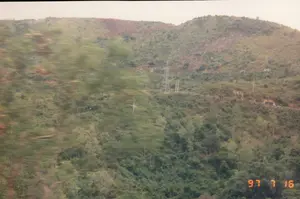 Dấu ngày tháng trên ảnh là '97 7 16'. Bức ảnh chụp phong cảnh một vùng đồi núi, có thể là ở khu vực Quảng Ngãi hoặc Bình Định. Cảnh vật dường như được chụp từ một phương tiện đang di chuyển, thể hiện qua hình ảnh cây cối ở tiền cảnh bị mờ nhòe do tốc độ. Phía sau là những ngọn đồi trập trùng được bao phủ bởi cây xanh. Trên sườn đồi có một cột điện cao thế và một con đường đất uốn lượn. Một chiếc xe, trông giống xe tải, đang chạy trên con đường này, gần một đoạn trông như đường hầm hoặc đường xẻ qua núi. Một mảng đất trống màu nâu đỏ xuất hiện trên đỉnh đồi, có thể là dấu vết của sạt lở hoặc hoạt động xây dựng.。（日本語訳）。写真の日付スタンプは「'97 7 16」です。この写真は、クアンガイ省またはビンディン省の丘陵地帯の風景を撮影したものです。この風景は移動中の乗り物から撮影されたようで、前景の木々が速度によってぼやけていることからそれが分かります。後方には、緑の木々に覆われた起伏のある丘が連なっています。丘の中腹には高圧電線の鉄塔と、曲がりくねった未舗装の道があります。丘の頂上には赤茶色の裸地が現れており、地滑りまたは建設活動の痕跡である可能性があります。