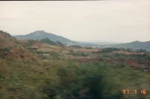 Bức ảnh này, có dấu ngày tháng '97 7 16' ở góc dưới bên phải, ghi lại một khung cảnh thiên nhiên rộng lớn ở vùng nông thôn. Đây là một cảnh quan đồi núi trập trùng. Ở phía trước và trung tâm, các ngọn đồi được bao phủ bởi cây cối và thảm thực vật xanh, xen kẽ với những mảng đất đỏ. Ở phía xa, có thể thấy những dãy núi cao hơn mờ ảo và một vùng thung lũng. Bầu trời u ám, không có nắng. Phần tiền cảnh bị nhòe do chuyển động, cho thấy bức ảnh có khả năng được chụp từ một phương tiện đang di chuyển trên đường.。（日本語訳）。この写真には、右下に「'97 7 16」の日付スタンプがあり、田舎の広大な自然の風景が記録されています。これは起伏のある丘陵地帯の風景です。手前と中央では、丘は木々や緑の植生に覆われ、赤土の区画が点在しています。遠くには、より高い山々がかすんで見え、谷間の地域が見えます。空は曇っており、太陽は出ていません。前景部分は動きによってぼやけており、写真が道路を移動中の乗り物から撮影された可能性が高いことを示しています。