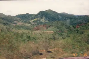 Có dấu ngày tháng '97 7 16' ở góc dưới bên phải của hình ảnh. Đây là quang cảnh một vùng đồi núi ở miền quê. Các ngọn đồi được bao phủ bởi cây cối và thảm thực vật xanh tươi. Trên sườn đồi, có những khoảng đất đã được phát quang, để lộ ra đất màu nâu đỏ, cho thấy dấu hiệu của hoạt động canh tác nông nghiệp, có thể là làm nương rẫy. Phần tiền cảnh của bức ảnh bị mờ, cho thấy nó có thể được chụp từ một phương tiện đang di chuyển. Bầu trời u ám và có nhiều mây.。（日本語訳）。画像の右下隅に「'97 7 16」という日付の印があります。これは田舎の丘陵地帯の光景です。丘は木々や青々とした植生に覆われています。丘の中腹には、開墾された土地があり、赤褐色の土が露出しており、おそらくは焼き畑農業のような農作業の痕跡が見られます。写真の前景はぼやけており、移動中の乗り物から撮影された可能性を示唆しています。空はどんよりとしており、雲が多いです。