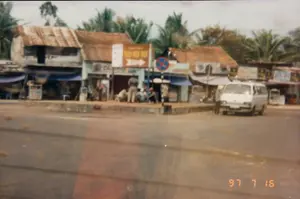 Bức ảnh chụp một khung cảnh ven đường ở một khu vực có vẻ là thị trấn hoặc vùng nông thôn ở Việt Nam vào ngày 16 tháng 7 năm 1997. Dọc bên đường là một dãy nhà cấp bốn, một số lợp mái ngói, một số lợp mái tôn đã cũ và gỉ sét. Các ngôi nhà này có vẻ là các cửa hàng hoặc nhà ở kết hợp kinh doanh, với mái hiên bằng bạt màu xanh được căng ra phía trước. Trên vỉa hè, một nhóm người đang tụ tập, một vài người đang ngồi xổm trong khi những người khác đứng, dường như đang trò chuyện hoặc chờ đợi. Một chiếc xe van màu trắng đang đỗ ở lề đường bên phải. Phía sau dãy nhà là những hàng cây dừa xanh tươi. Có một số biển báo: một biển báo giao thông hình tròn, viền đỏ, nền xanh có gạch chéo đỏ (biển báo cấm dừng và đỗ xe). Bên cạnh đó là một biển chỉ dẫn hình chữ nhật màu nâu cam với chữ trắng và một mũi tên chỉ sang phải, nhưng chữ viết trên đó rất mờ và không thể đọc được. Các biển hiệu cửa hàng khác cũng bị mờ. Trên tường của một ngôi nhà có gắn hai cục nóng máy lạnh. Bức ảnh có vẻ được chụp từ bên trong một phương tiện đang di chuyển.。（日本語訳）。1997年7月16日に撮影された、ベトナムの町か田舎と思われる地域の道端の風景写真です。道沿いには平屋の家々が並んでおり、一部は瓦屋根、一部は古びて錆びたトタン屋根です。これらの家は店舗または店舗兼住宅のようで、青い防水シートの日よけが前に張り出されています。歩道では一群の人々が集まっており、何人かはしゃがみ込み、他の人々は立っていて、雑談しているか何かを待っているようです。白いバンが道路の右側に駐車しています。家々の後ろには、青々としたヤシの木が並んでいます。いくつかの標識があります。一つは円形の交通標識で、赤い縁取り、青い背景に赤い斜線が入っています（駐停車禁止の標識）。その隣には、オレンジがかった茶色に白い文字と右を指す矢印がある長方形の案内標識がありますが、そこに書かれている文字は非常にぼやけていて読むことができません。他の店の看板も同様にぼやけています。ある家の壁には、エアコンの室外機が2台設置されています。この写真は、移動中の乗り物の中から撮影されたようです。