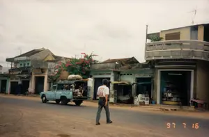 Bối cảnh là một con đường tại một thị trấn ở Việt Nam, được chụp vào ngày 16 tháng 7 năm 1997. Dọc theo con đường là những ngôi nhà phố liền kề, có cả nhà một tầng và hai tầng, với kiến trúc đặc trưng của thời kỳ đó. Một số ngôi nhà trông đã cũ và xuống cấp, trong khi một ngôi nhà bên phải có ban công bằng bê tông trông hiện đại hơn. Một giàn hoa giấy lớn màu hồng đang nở rộ phía trước một ngôi nhà. Trên đường, một chiếc xe tải nhỏ màu xanh lam đang đỗ lại. Trên thùng xe có nhiều người đang ngồi cùng với các bao tải hàng hóa. Một người đàn ông đang đứng ở phía sau xe. Ở giữa đường, một người đàn ông mặc áo phông trắng và quần dài đang đi bộ. Ven đường, có các cửa hàng đang hoạt động. Cửa hàng ở ngoài cùng bên phải có cửa mở rộng, bên trong có các kệ trưng bày hàng hóa, cho thấy đây có thể là một tiệm tạp hóa. Phía trước một cửa hàng khác, một người phụ nữ đội nón lá đang ngồi. Có một vài biển hiệu trên các tòa nhà nhưng chữ đã mờ, không thể đọc rõ nội dung.。（日本語訳）。背景は1997年7月16日に撮影された、ベトナムのある町の通りです。通り沿いには、平屋と二階建ての両方がある、その時代特有の建築様式のタウンハウスが隣接して建ち並んでいます。いくつかの家は古く、老朽化しているように見えますが、右側にある家はコンクリートのバルコニーがあり、より現代的に見えます。ある家の前では、大きなピンク色のブーゲンビリアの棚が満開です。路上には、青い小型トラックが一台停まっています。荷台には、多くの人々が荷物の入った袋と一緒に座っています。一人の男性が車の後方に立っています。道路の中央では、白いTシャツと長ズボンを履いた男性が歩いています。道端では、いくつかの店が営業しています。一番右端の店はドアが大きく開いており、中には商品を陳列する棚が見え、ここが食料雑貨店である可能性を示しています。別の店の前では、ノンラーをかぶった女性が座っています。