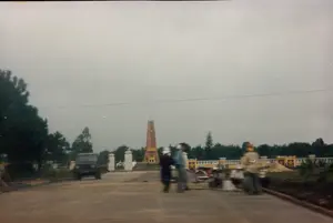 Bức ảnh chụp một khung cảnh bên đường vào một ngày trời u ám, có thể ở khu vực Quảng Ngãi hoặc Bình Định. Ở phía sau, nổi bật là một đài tưởng niệm hình tháp, màu nâu đỏ, trên đỉnh có gắn ngôi sao vàng năm cánh. Có một tấm biển trên đài tưởng niệm nhưng chữ viết quá mờ để đọc. Lối vào khu tưởng niệm có hai cột trụ màu trắng. Dọc theo khu vực là một hàng rào sơn màu trắng và vàng. Trên con đường nhựa ở phía trước, một chiếc xe ô tô kiểu quân sự (giống xe jeep) đang ở bên trái. Ở giữa và bên phải đường, một nhóm người đang tụ tập. Một số người đứng, một số ngồi xổm bên cạnh vài chiếc xe máy đang đậu. Hoạt động của họ không rõ ràng nhưng có vẻ họ đang dừng chân nghỉ ngơi bên đường.。（日本語訳）。写真は、おそらくクアンガイ省かビンディン省の地域で、曇りの日の道端の風景を撮影したものです。後方には、赤褐色の塔の形をした記念碑が際立っており、その頂上には五芒星の黄色い星が取り付けられています。記念碑には銘板がありますが、文字がぼやけていて読めません。記念碑エリアへの入り口には、2本の白い柱があります。そのエリアに沿って、白と黄色に塗られた柵があります。手前のアスファルトの道路上、左側には軍用車風の自動車（ジープのような）が1台あります。道路の中央と右側には、人々の集団が集まっています。何人かは立っており、何人かは駐車している数台のバイクの隣でしゃがんでいます。彼らの活動は明確ではありませんが、道端で休憩しているように見えます。