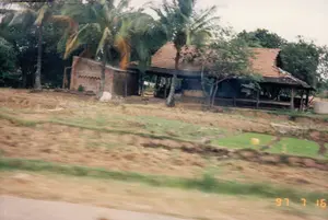 Bức ảnh chụp một khung cảnh nông thôn ở Việt Nam, có thể là ở khu vực Quảng Ngãi hoặc Bình Định, vào ngày 16 tháng 7 năm 1997, như được ghi nhận bởi dấu ngày tháng '97 7 16' ở góc dưới bên phải. Phong cảnh cho thấy một ngôi nhà mái ngói đỏ, một phần mái có vẻ đã bị hư hỏng. Bên cạnh ngôi nhà chính là một công trình phụ xây bằng gạch đỏ chưa hoàn thiện. Xung quanh là rất nhiều cây cối, đặc biệt là những cây dừa cao. Phía trước ngôi nhà, trên một sườn dốc, là một thửa ruộng nhỏ trồng mạ non xanh mướt, cho thấy hoạt động canh tác nông nghiệp. Toàn bộ bức ảnh có vẻ được chụp từ một phương tiện đang di chuyển, vì phần tiền cảnh bị mờ nhòe. Không có người nào được nhìn thấy rõ trong ảnh.。（日本語訳）。写真は、右下隅にある「97 7 16」という日付スタンプに記録されているように、1997年7月16日に、ベトナムのクアンガイ省またはビンディン省の地域で撮影された田舎の風景を写している。母屋の隣には、未完成の赤レンガ造りの付属の建物がある。周りにはたくさんの木々、特に高いココヤシの木がある。家の前、坂の上には、青々とした若い稲が植えられた小さな田んぼがあり、農業活動が行われていることを示している。