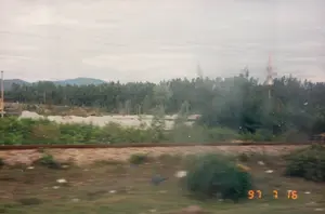 Bức ảnh ghi lại cảnh quan nông thôn được chụp từ một phương tiện đang di chuyển, có thể là tàu hỏa, vào ngày 16 tháng 7 năm 1997. Ở tiền cảnh là đường ray xe lửa. Phía sau đường ray là một khu vực có vẻ như là bãi tập kết vật liệu xây dựng hoặc khai thác khoáng sản, với những đống lớn vật liệu màu trắng (có thể là cát hoặc đá). Một rừng cây rậm rạp mọc phía sau khu vực này. Ở phía xa bên phải, một công trình kiến trúc cao, có thể là một đài tưởng niệm, vươn lên trên những ngọn cây. Phía sau cùng là dãy núi mờ ảo dưới bầu trời nhiều mây. Có các đường dây điện chạy ngang qua khung cảnh. Dấu ngày tháng "97 7 16" được in ở góc dưới bên phải của bức ảnh.。（日本語訳）。1997年7月16日に、移動中の乗り物、おそらくは列車から撮影された田園風景を記録した写真です。前景には線路があります。このエリアの後ろには、鬱蒼とした森が広がっています。遠くの右側には、高い建築物、おそらくは記念碑が、木々の梢の上にそびえ立っています。一番奥には、曇り空の下にぼんやりとした山脈が見えます。電線が風景を横切っています。写真の右下隅には「97 7 16」という日付が印字されています。