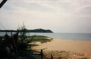 Bức ảnh ghi lại một khung cảnh ven biển yên bình, được chụp vào ngày 16 tháng 7 năm 1997, như được thể hiện bởi dấu ngày tháng "97 7 16" ở góc dưới bên phải. Địa điểm là một bãi biển hoang sơ với dải cát vàng. Mặt biển lặng và ở phía xa có một hòn đảo hoặc bán đảo được cây cối bao phủ. Bầu trời có mây trắng. Ở tiền cảnh bên trái, có một hàng rào gỗ đơn sơ và một phần của một công trình tạm bợ, có thể là một cái chòi của ngư dân, được dựng bằng các vật liệu tự nhiên như tre, nứa. Một vài cây phi lao và thảm thực vật ven biển mọc trên cát. Cảnh vật vắng lặng, không có hoạt động nào của con người được ghi lại, mang lại cảm giác về một vùng quê ven biển tĩnh lặng và chưa bị tác động nhiều.。（日本語訳）。この写真は、穏やかな海辺の風景を記録したもので、右下の「97 7 16」という日付スタンプが示すように、1997年7月16日に撮影されました。場所は、金色の砂が広がる手付かずのビーチです。海は穏やかで、遠くには木々に覆われた島、あるいは半島が見えます。空には白い雲が浮かんでいます。左の前景には、簡素な木製の柵と、竹などの自然素材で建てられた、おそらく漁師小屋と思われる仮設の建物の一部があります。砂の上には、数本のモクマオウと海辺の植生が生えています。風景は静かで、人の活動は見られず、静かで手付かずの海辺の田舎という印象を与えます。