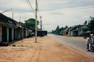 Bối cảnh là một con đường nhựa ở một vùng quê hoặc thị trấn nhỏ ở Việt Nam vào năm 1997. Lề đường là đất cát.。Bên trái là một dãy nhà cấp bốn liền kề, trông cũ kỹ, có thể là nhà ở hoặc cửa hàng. Một chiếc xe tải màu xanh đậm đang đậu bên lề đường. Gần chiếc xe tải có một chiếc ô màu xanh lá cây, có thể là một quán nước nhỏ ven đường.。Bên phải đường, hai người đàn ông đang đi trên một chiếc xe máy. Phía xa hơn là những ngôi nhà khác và cây cối, bao gồm cả cây dừa. Hệ thống cột điện và dây điện chằng chịt bắc ngang qua đường. Bầu trời có mây.。Trên một ngôi nhà ở phía xa bên phải, có một tấm biển nhưng chữ viết đã bị mờ và không thể đọc rõ. Không có văn bản nào khác có thể đọc được.。（日本語訳）。舞台は1997年、ベトナムの田舎、または小さな町のアスファルト道路です。路肩は砂地です。濃い青色のトラックが路肩に駐車しています。トラックの近くには緑色の傘があり、道端の小さな飲み物屋台かもしれません。道路の右側では、男性2人がバイクに乗っています。さらに遠くには、ココヤシの木を含む他の家々や木々があります。電柱と電線が複雑に絡み合って道を横切っています。空には雲があります。右奥の家には看板がありますが、文字は薄れていてはっきりと読むことはできません。他に読み取れる文字はありません。
