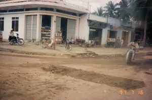 Bối cảnh là một con đường đất tại một thị trấn hoặc làng quê ở Việt Nam, chụp vào ngày 16 tháng 7 năm 1997. Bên trái là một ngôi nhà hai tầng kiên cố, tầng trệt được sử dụng làm cửa hàng. Ngôi nhà có cửa cuốn bằng kim loại đang mở, để lộ các kệ hàng hóa bên trong, trông giống một tiệm tạp hóa. Trên tường có gắn số nhà "222". Bên cạnh là một dãy các cửa hàng cấp bốn nhỏ hơn. Một trong số đó có biển hiệu với chữ "PHÚ" có thể đọc được, các biển hiệu khác bị mờ và không thể đọc rõ. Con đường không được trải nhựa, bề mặt là đất và gồ ghề. Có một số người và phương tiện đi lại. Một người đàn ông đội mũ đang ngồi trên chiếc xe máy kiểu Honda Cub đậu bên đường. Một người khác đang điều khiển một chiếc xe tay ga (scooter) đang di chuyển. Một vài chiếc xe máy và xe đạp khác đang đậu trước các cửa hàng. Phía sau các tòa nhà là những hàng cây dừa, một hình ảnh đặc trưng của làng quê Việt Nam.。（日本語訳）。背景はベトナムの町または田舎にある未舗装の道で、1997年7月16日に撮影されたものです。左側には頑丈な2階建ての家があり、1階は店舗として使用されています。その家には金属製のシャッターがあり、開いていて、中の商品棚が見え、雑貨店のように見えます。壁には番地の「222」が取り付けられています。その隣には、より小さな平屋建ての店が並んでいます。そのうちの一つには「PHÚ」という文字が読める看板があり、他の看板はぼやけていてはっきりと読むことはできません。道は舗装されておらず、地面は土ででこぼこです。数人の人々と乗り物が行き来しています。帽子をかぶった男性が、道端に停められたホンダ・カブ型のバイクに座っています。別の人が移動中のスクーターを運転しています。他の数台のバイクと自転車が店の前に停められています。建物の後ろにはココナッツの木が並んでおり、ベトナムの田舎の典型的な光景です。
