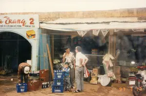 Bối cảnh là phía trước một cửa hàng tạp hóa hoặc một cơ sở kinh doanh dịch vụ ở Việt Nam vào năm 1997. Nền đất ẩm ướt và lầy lội. Trên tấm biển hiệu lớn có chữ "...a Trang 2" và dòng chữ nhỏ hơn bên dưới: "THUÊ ĐỒ CƯỚI, BÀN GHẾ, PHÔNG MÀN, BÁT ĐĨA...". Điều này cho thấy cửa hàng cung cấp dịch vụ cho thuê đồ dùng cho đám cưới và các sự kiện. Một người đàn ông mặc áo khoác nâu đang cúi xuống sắp xếp các chai nước vào két nhựa màu xanh của hãng "Vĩnh hảo". Gần đó, hai người đàn ông và một cậu bé đang đứng nói chuyện. Bên trong cửa hàng, hàng hóa được bày trên kệ. Phía trước có nhiều két nước giải khát bằng chai thủy tinh (như Pepsi, Vĩnh hảo), các thùng hàng carton (có thể thấy hộp bột giặt Tide), và một chiếc mâm nhôm. Một chiếc xe máy kiểu cũ đang đậu ở bên phải.。（日本語訳）。1997年のベトナムにある雑貨店またはサービス業の店の前が舞台です。地面は湿っていてぬかるんでいます。大きな看板には「...a Trang 2」という文字と、その下に小さな文字で「結婚式用品、テーブル・椅子、背景幕、食器…のレンタル」と書かれています。これは、この店が結婚式やイベント用品のレンタルサービスを提供していることを示しています。茶色いジャケットを着た男性が、かがんで「Vĩnh hảo」社の青いプラスチックケースに水のボトルを並べています。近くでは、男性2人と少年1人が立って話しています。店内では、商品が棚に陳列されています。右側には旧式のバイクが1台停まっています。