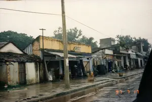 Bức ảnh ghi lại khung cảnh một con phố ở Việt Nam vào một ngày mưa, chụp ngày 17 tháng 7 năm 1997. Con đường nhựa ướt sũng, có nhiều vũng nước, phản chiếu bầu trời xám xịt. Dọc bên đường là một dãy nhà phố liền kề, chủ yếu là các cửa hàng kinh doanh. Kiến trúc các ngôi nhà khá đa dạng, từ nhà cấp bốn lợp ngói cũ kỹ, tường vôi đã bạc màu ở bên trái, đến những ngôi nhà tầng kiên cố hơn. Một cửa hàng có mặt tiền sơn vàng nổi bật với tấm biển hiệu có thể đọc được chữ "DENSO", cho thấy đây có thể là một tiệm bán phụ tùng hoặc sửa chữa ô tô, xe máy. Phía trước các cửa hàng có mái hiên bằng tôn hoặc bạt để che mưa nắng. Bên lề đường có một chồng lốp xe, một chiếc xe đạp, và vài chiếc thùng gỗ. Một cột điện bê tông lớn đứng ở vị trí trung tâm, cùng với hệ thống dây điện chằng chịt phía trên. Có một vài người dân đang hoạt động gần các cửa hàng, nhưng không khí chung khá yên ắng, có thể do trời mưa. Góc dưới bên phải ảnh có dấu ngày tháng màu cam "97 7 17".。（日本語訳）。この写真は1997年7月17日に撮影された、雨の日のベトナムのある通りの風景を記録したものです。アスファルトの道はびしょ濡れで、多くの水たまりがあり、灰色の空を反射しています。道沿いには商店が連なる長屋が並んでおり、そのほとんどが商業店舗です。家々の建築様式は非常に多様で、左側にある古い瓦屋根で漆喰の壁が色褪せた平屋から、より頑丈な階建ての家まであります。ある店は黄色く塗られたファサードが際立っており、「DENSO」という文字が読み取れる看板があり、ここが自動車やバイクの部品販売店または修理店である可能性を示しています。店の前には、雨や日差しを避けるためのトタンや防水シートでできた庇があります。道端には積み重ねられたタイヤ、一台の自転車、そしていくつかの木箱があります。大きなコンクリートの電柱が中央に立っており、その上には複雑に絡み合った電線があります。店の近くで活動している住民が数人いますが、全体の雰囲気はかなり静かで、雨が降っているためだと思われます。写真の右下隅には、「97 7 17」というオレンジ色の年月のスタンプがあります。