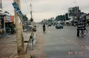 Bối cảnh là một con đường tại một đô thị ở Việt Nam, được chụp vào ngày 17 tháng 7 năm 1997. Con đường được trải nhựa nhưng hai bên lề đường còn là đất, lầy lội và có những vũng nước, có thể là sau một cơn mưa.。Về chữ viết, ở phía trước bên trái có một cột điện bê tông, trên đó gắn một biển báo tên đường màu xanh bị gãy. Biển báo ghi tên hai con đường: 'ĐƯỜNG LÊ LỢI' và 'ĐƯỜNG PHAN.Đ.PHUNG' (có thể là đường Phan Đình Phùng). Trên cột điện còn có các ký hiệu 'BT2' và '10'. Ở phía xa bên phải, có các biển hiệu cửa hàng, trong đó có thể đọc được một phần chữ 'PHƯƠNG' và 'TUANTR...'.。Hoạt động trên đường phố khá tấp nập. Nhiều người đang di chuyển bằng các phương tiện phổ biến thời đó như xe đạp và xe máy. Một người đàn ông đang đạp xe trên đường. Ở phía bên phải, một người đội nón lá đang đẩy một chiếc xe đạp chở hai vật hình trụ lớn, cồng kềnh. Ngoài ra còn có một vài chiếc ô tô kiểu cũ đang lưu thông. Dọc hai bên đường là những dãy nhà phố thấp tầng, vừa để ở vừa kinh doanh, với nhiều biển hiệu. Hệ thống cột điện và dây điện chằng chịt là một hình ảnh đặc trưng của thời kỳ này.。（日本語訳）。背景はベトナムのある都市の道路で、1997年7月17日に撮影されたものです。道路はアスファルトで舗装されていますが、両側の路肩はまだ土で、ぬかるんで水たまりができており、雨が降った後かもしれません。文字については、左手前にコンクリートの電柱があり、そこには壊れた青い道路標識が取り付けられています。標識には「ĐƯỜNG LÊ LỢI（レ・ロイ通り）」と「ĐƯỜNG PHAN.Đ.PHUNG（ファン・ディン・フン通りである可能性あり）」という2つの通りの名前が書かれています。電柱には「BT2」と「10」という記号もあります。右奥には店の看板があり、その中には「PHƯƠNG」と「TUANTR...」という文字の一部が読み取れます。通りでの活動はかなり賑やかです。多くの人々が、自転車やバイクといった当時一般的だった乗り物で移動しています。一人の男性が道路で自転車を漕いでいます。右側では、ノンラー（菅笠）をかぶった人が、大きくてかさばる2つの円筒形の物を積んだ自転車を押しています。その他にも、数台の旧式の自動車が通行しています。道路の両脇には、住居兼店舗として使われている低層のタウンハウスが、多くの看板を掲げて立ち並んでいます。張り巡らされた電柱と電線のシステムは、この時代の特徴的な光景です。