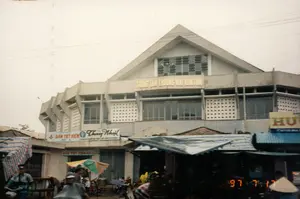 Bức ảnh chụp mặt tiền Trung tâm Thương mại Kontum và khu chợ nhộn nhịp phía trước vào ngày 17 tháng 7 năm 1997. Tòa nhà trung tâm thương mại là một công trình bê tông lớn với kiến trúc mái tam giác, trên đó có dòng chữ "TRUNG TÂM THƯƠNG MẠI KONTUM".。Phía dưới là cảnh sinh hoạt và buôn bán tấp nập. Nhiều quầy hàng được dựng lên với mái che bằng bạt. Người dân đi lại bằng xe máy và xe đạp, một số người mặc áo mưa, và một người phụ nữ đội nón lá. Các biển hiệu có thể đọc được bao gồm cửa hàng "Thủy Nhựt" và một điểm dịch vụ "BÀN TIẾT KIỆM SỐ 1", cho thấy đây là một khu vực kinh doanh đa dạng. Khung cảnh phản ánh không khí thương mại sôi động của thành phố Kontum vào thời điểm đó.。（日本語訳）。1997年7月17日に撮影された、コントゥム商業センターの正面とその前の賑やかな市場の写真。商業センターの建物は、三角形の屋根を持つ大きなコンクリートの建造物で、その上には「TRUNG TÂM THƯƠNG MẠI KONTUM」という文字が書かれています。その下には、賑やかな生活と商売の光景が広がっています。多くの露店が防水シートの屋根で建てられています。人々はバイクや自転車で往来し、何人かはレインコートを着ており、一人の女性はノンラーをかぶっています。読み取れる看板には「Thủy Nhựt」店や「BÀN TIẾT KIỆM SỐ 1」というサービス拠点が含まれており、ここが多様な商業地域であることを示しています。この光景は、当時のコントゥム市の活気ある商業の雰囲気を反映しています。