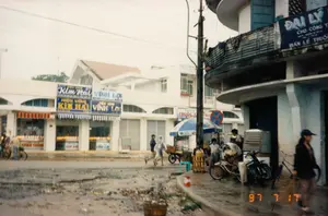 Đây là hình ảnh một góc phố tại Việt Nam, được chụp vào ngày 17 tháng 7 năm 1997. Khung cảnh cho thấy một con đường nhựa ẩm ướt, có lẽ trời vừa mưa hoặc đang mưa nhỏ.。**Chữ viết:**。*   Trên dãy nhà phố màu trắng bên trái, có nhiều biển hiệu của các cửa hàng kinh doanh vàng bạc: "HIỆU VÀNG Kim Hải" và "TIỆM VÀNG VĨNH LỢI". Một biển hiệu khác có chữ "ĐÔNG THÀNH" nhưng bị che khuất.。*   Trên tòa nhà ở góc đường bên phải, có một biển hiệu lớn màu xanh: "ĐẠI LÝ ... CHO CÔNG T... BÁN LẺ THUỐC...", cho thấy đây có thể là một đại lý bán lẻ dược phẩm.。*   Ở phía sau, có một biển hiệu khác ghi "THUỐC T..." và một biển báo giao thông hình tròn màu xanh có gạch chéo đỏ (biển báo cấm dừng và đỗ xe).。**Bối cảnh và Hoạt động:**。*   **Địa điểm:** Một góc phố thương mại tại một đô thị. Sự hiện diện của nhiều tiệm vàng và một đại lý thuốc cho thấy đây là một khu vực buôn bán sầm uất.。*   **Hoạt động:** Cuộc sống thường nhật đang diễn ra. Một người mặc áo mưa ni lông đang đi bộ và đẩy một chiếc xe kéo tay. Những người khác đang đi lại trên vỉa hè và lòng đường. Một nhóm người đang tụ tập ở vỉa hè bên phải, gần một chiếc xe đạp và một chiếc xe máy.。*   **Vật thể:** Các tòa nhà là nhà ống nhiều tầng điển hình. Phương tiện giao thông trên đường gồm có xe đạp, xe máy và xe kéo tay. Một cây cột điện lớn với hệ thống dây điện chằng chịt đứng ở vị trí trung tâm. Một chiếc ô (dù) lớn màu xanh trắng được mở trên vỉa hè, có thể là của một người bán hàng rong. Trên ban công của tòa nhà bên phải có phơi quần áo.。（日本語訳）。これは1997年7月17日に撮影された、ベトナムのある街角の写真です。景色は湿ったアスファルトの道を示しており、おそらく雨が降った直後か、小雨が降っているのでしょう。**文字：**。*   左側の白いタウンハウスの列には、多くの金銀を商う店の看板があります：「HIỆU VÀNG Kim Hải」と「TIỆM VÀNG VĨNH LỢI」。別の看板には「ĐÔNG THÀNH」という文字がありますが、隠されています。*   右側の角の建物には、「ĐẠI LÝ ... CHO CÔNG T... BÁN LẺ THUỐC...」と書かれた大きな青い看板があり、これが医薬品の小売代理店である可能性を示しています。*   後方には、「THUỐC T...」と書かれた別の看板と、赤い斜線が入った青い円形の交通標識（駐停車禁止の標識）があります。**背景と活動：**。*   **場所：** ある都市の商業地区の街角。多くの金行と薬の代理店の存在は、ここが活気のある商業地域であることを示しています。*   **活動：** 日常の生活が繰り広げられています。ビニールのレインコートを着た人が歩きながらリヤカーを押しています。他の人々は歩道や車道を行き来しています。右側の歩道では、自転車とバイクの近くに人々が集まっています。*   **物体：** 建物は典型的な多層のチューブハウスです。道路上の交通手段には、自転車、バイク、リヤカーが含まれます。複雑に絡み合った電線を持つ大きな電柱が中央に立っています。歩道には青と白の大きな傘が開かれており、それは露天商のものかもしれません。右側の建物のバルコニーには洗濯物が干されています。