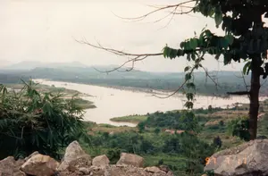 Bức ảnh ghi lại một khung cảnh thiên nhiên rộng lớn tại vùng cao nguyên, có khả năng là khu vực sông Sê San gần công trình thủy điện Yaly ở Kon Tum, được chụp vào ngày 17 tháng 7 năm 1997. Dấu ngày tháng '97 7 17' được in màu cam trên một tảng đá ở góc dưới bên phải.。Từ một vị trí trên cao, tầm nhìn bao quát một con sông lớn với dòng nước màu nâu đục, uốn lượn qua những ngọn đồi và núi non được bao phủ bởi cây cối xanh tươi. Ở tiền cảnh, có những tảng đá và một bụi tre ở bên trái, cùng với một cái cây ở bên phải. Dọc theo bờ sông, có những khu vực đất trống và một vài cấu trúc không rõ ràng hoặc cây khô chết đứng giữa dòng, có thể là dấu hiệu của hoạt động xây dựng hoặc sự thay đổi của hệ sinh thái do dự án thủy điện gây ra. Bầu trời u ám, không có hoạt động cụ thể nào của con người được ghi lại, chủ yếu là một cảnh quan tĩnh lặng.。（日本語訳）。この写真は、1997年7月17日に撮影された、高原地帯の広大な自然風景を記録したもので、おそらくコントゥム省のヤリー水力発電所近くのセサン川地域だと思われます。右下の角にある岩の上には、「'97 7 17」という日付がオレンジ色で印字されています。高い場所から見ると、青々とした木々に覆われた丘や山々の間を、濁った茶色の大きな川が蛇行しているのが一望できます。前景には、左側にいくつかの岩と竹やぶがあり、右側には一本の木があります。川岸に沿って、空き地やいくつかの不明瞭な構造物、あるいは流れの中に立ち枯れた木があり、これらは建設活動の兆候か、水力発電プロジェクトによって引き起こされた生態系の変化である可能性があります。空は曇っており、人間の具体的な活動は記録されておらず、主に静寂な景観です。