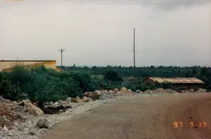 写真には右下隅にオレンジ色の日付スタンプがあります：「\'97 7 17」。これは、ある農村地域の土の道または砂利道の光景で、テイグエン地方である可能性があります。道は右にカーブしており、路肩は大きな石と砂利で固められています。道の両脇には雑草や茂みが生い茂っています。左側には、黄色い壁の建築物の一部があります。さらに奥には、平屋建ての家か長い小屋があり、簡素な屋根で覆われています。写真には2本の柱があります。1本は木製の電柱で、もう1本にはアンテナが取り付けられています。1本の細い電線が上方を横切っています。茂みの後ろに、青い自動車の一部が見えます。遠くには、曇り空の下、幾重にも連なる丘や山の景色が広がっています