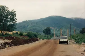 Bối cảnh là một con đường đất đỏ gồ ghề ở một vùng đồi núi. Một chiếc ô tô màu trắng kiểu cũ đang ở trên đường, hướng về phía xa. Con đường có vẻ như đang được xây dựng hoặc sửa chữa, với đất và đá chất thành đống ở ven đường bên trái. Phông nền là một dãy núi rộng lớn, cây cối rậm rạp bao phủ. Đỉnh núi xa xa chìm trong sương mù hoặc mây. Trên sườn núi có những mảng đất trống màu nâu, có thể là dấu hiệu của hoạt động xây dựng. Dọc đường đi có thể thấy các cột điện, bao gồm cả những trụ bê tông lớn, cho thấy đây có thể là khu vực gần một công trường xây dựng cơ sở hạ tầng quan trọng. Bầu trời u ám, không có nắng.。（日本語訳）。背景は、丘陵地帯にあるでこぼこの赤土の道です。一台の旧式の白い車が道路上にあり、遠くに向かっています。その道は建設中か修理中のようで、左側の道端には土や石が山積みにされています。背景は、木々が鬱蒼と茂る広大な山脈です。遠くの山頂は霧か雲に沈んでいます。山の斜面には茶色い裸地があり、それは建設活動の兆候かもしれません。道沿いには、大きなコンクリート柱も含む電柱が見られ、ここが重要なインフラ建設現場の近くである可能性を示しています。空は曇っており、日差しはありません。