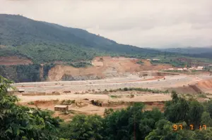 Bức ảnh ghi lại quang cảnh một đại công trường xây dựng, có thể là công trình thủy điện Yali ở Kon Tum, vào ngày 17 tháng 7 năm 1997, như được chỉ ra bởi dấu ngày tháng '97 7 17' màu vàng ở góc dưới bên phải. Khung cảnh cho thấy một thung lũng rộng lớn đang trong quá trình thi công. Một con đập lớn bằng đất và đá đang được xây dựng, với nhiều khu vực đất đã bị đào xới, để lộ ra lớp đất đỏ. Trên công trường có nhiều phương tiện cơ giới hạng nặng như máy ủi và xe tải đang hoạt động. Rải rác trong khu vực là các công trình tạm như lán trại cho công nhân. Bao quanh công trường là những ngọn đồi và núi được bao phủ bởi rừng cây xanh rậm rạp dưới bầu trời u ám, nhiều mây.。（日本語訳）。この写真は、右下の黄色い日付スタンプ「'97 7 17」が示すように、1997年7月17日の、おそらくコントゥム省のヤーリー水力発電所プロジェクトである大規模な建設現場の光景を記録したものです。その光景は、広大な渓谷が工事中であることを示しています。土と石でできた大きなダムが建設されており、多くの土地が掘削され、赤い土の層が露出しています。建設現場では、ブルドーザーやトラックなどの多くの重機が稼働しています。この地域には、作業員用の仮設小屋のような一時的な建物が点在しています。建設現場の周りは、曇り空の下、緑豊かな森に覆われた丘や山々が囲んでいます。