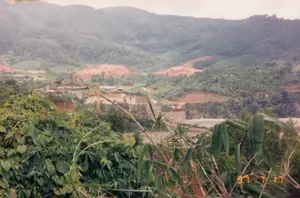 Trong ảnh có dấu ngày tháng màu cam ở góc dưới bên phải ghi "97 7 17".。Bức ảnh chụp toàn cảnh một công trường xây dựng quy mô lớn, có khả năng là công trình đập thủy điện Yaly ở vùng Kon Tum, được chụp từ một vị trí trên cao. Hoạt động chính đang diễn ra là việc xây dựng một con đập lớn. Ở trung tâm bức ảnh, có thể thấy các kết cấu bê tông lớn của thân đập đang trong quá trình thi công, cùng với ít nhất một chiếc cần cẩu tháp màu vàng. Xung quanh công trình là những khu vực đất đỏ rộng lớn đã được san ủi và đào bới.。Các vật thể trong ảnh bao gồm:。- Tiền cảnh: Cây cối, bụi rậm và tre nứa mọc um tùm.。- Trung cảnh: Công trường xây dựng với đập bê tông, cần cẩu, và một vài tòa nhà nhỏ có thể là lán trại công nhân.。- Hậu cảnh: Những dãy đồi núi trập trùng được bao phủ bởi rừng cây xanh rậm rạp. Một số sườn đồi cũng có những mảng đất trống màu đỏ do bị phát quang.。（日本語訳）。写真の右下隅には、「97 7 17」と記されたオレンジ色の日付スタンプがあります。写真は、高所から撮影された、コントゥム地方のヤーリー水力発電ダム事業である可能性のある大規模な建設現場の全景を写しています。行われている主な活動は、大きなダムの建設です。写真の中央では、建設中のダム本体の大きなコンクリート構造物と、少なくとも1基の黄色いタワークレーンが見られます。工事現場の周囲には、整地され掘削された広大な赤土の区域があります。写真内の物体は以下の通りです：。- 前景：生い茂る木々、茂み、竹。- 中景：コンクリートのダム、クレーン、そして作業員の宿舎である可能性のあるいくつかの小さな建物がある建設現場。- 背景：鬱蒼とした緑の森に覆われた起伏のある丘や山々。一部の丘の斜面には、伐採によってできた赤土のむき出しの土地も見られます。