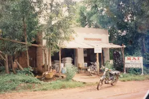 Trên bức ảnh có một bảng hiệu lớn màu trắng với chữ đỏ và xanh, nội dung: 'MAI - ĐẠI LÝ - CHUYÊN DOANH GẠO THỨC ĂN GIA SÚC - BÁN SỈ VÀ LẺ - GIÁ PHẢI CHĂNG'. Có một bảng hiệu nhỏ khác treo dưới mái hiên nhưng chữ viết không thể đọc được. Dưới góc phải bức ảnh có dấu ngày tháng '97 7 17'.。Bối cảnh là một đại lý nhỏ ven một con đường đất đỏ ở vùng nông thôn. Cửa hàng là một ngôi nhà cấp bốn, tường trát xi măng và có một phần tường gạch chưa trát. Phía trước có mái hiên tạm được dựng lên. Bên trong và trước hiên nhà có nhiều bao tải lớn màu trắng được xếp chồng lên nhau, cho thấy đây là nơi bán các mặt hàng như gạo và thức ăn gia súc. Có hai chiếc xe máy kiểu cũ, có thể là dòng xe Honda Cub, một chiếc đậu bên trong hiên và một chiếc đậu ở lề đường. Một người đang ngồi trên băng ghế gỗ bên trong cửa hàng. Cảnh vật xung quanh có nhiều cây cối xanh tươi.。（日本語訳）。写真には、赤と青の文字が書かれた大きな白い看板があり、その内容は「マイ - 代理店 - 米・家畜飼料専門 - 卸売・小売 - お手頃価格」です。軒下には別の小さな看板が掛かっていますが、文字は読めません。写真の右下には「97 7 17」という日付の印があります。背景は、田舎の赤土の道の脇にある小さな代理店です。店は平屋建ての家で、壁はセメントで塗られており、一部にまだ塗られていないレンガの壁があります。正面には仮設の軒が建てられています。家の中と軒先には、大きな白い袋がたくさん積み重ねられており、ここが米や家畜飼料のような商品を販売する場所であることを示しています。古い型のバイクが2台あり、おそらくホンダ・カブの車種で、1台は軒の内側に、もう1台は道端に停められています。店内で一人の人が木製のベンチに座っています。周囲の景色には青々とした木々がたくさんあります。