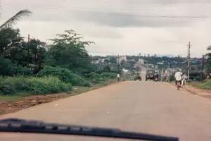 Bức ảnh chụp một con đường nhựa ở khu vực nông thôn hoặc ngoại ô, có thể là ở vùng lân cận Kon Tum, nhìn từ bên trong một chiếc xe hơi qua kính chắn gió. Con đường trải dài về phía xa, hướng lên một ngọn đồi nơi có các tòa nhà của một thị trấn hoặc khu dân cư. Hai bên đường là cây cối và bụi rậm xanh tươi, cùng với các cột điện. Trên đường, có nhiều người dân địa phương đang di chuyển. Một người đàn ông mặc áo trắng đang đi xe đạp, và những người khác cũng đang đi xe đạp và xe máy. Một chiếc xe jeep và một phương tiện trông giống máy cày đang chạy ở phía trước. Một người đang đi bộ bên lề đường. Khung cảnh thể hiện hoạt động giao thông và sinh hoạt thường ngày vào một ngày trời nhiều mây năm 1997. Không có văn bản hay biển hiệu nào có thể đọc được trong ảnh.。（日本語訳）。写真は、田舎または郊外のアスファルトの道を、おそらくコントゥム近郊で、車内からフロントガラス越しに撮影したものです。道は遠くまで伸びており、町または住宅地の建物がある丘に向かっています。道の両側には青々とした木々や茂みがあり、電柱も立っています。道路上では、多くの地元の人々が移動しています。白いシャツを着た男性が自転車に乗っており、他の人々も自転車やバイクに乗っています。前方では、一台のジープとトラクターのような乗り物が走っています。一人の人が路肩を歩いています。この光景は、1997年のある曇りの日の、日常的な交通と生活の様子を表しています。写真の中には、読み取れる文字や看板はありません。