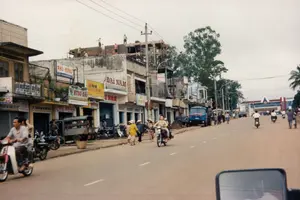 Các chữ viết trên các biển hiệu có thể nhận dạng bao gồm: PHÚ HƯNG, HƯNG HẢI, ĐẠI NAM, Kodak EXPRESS, và Cafe GIẢI KHÁT. Ngoài ra còn có các biển hiệu khác bị che khuất hoặc mờ không đọc được, như một biển hiệu có chữ "...QUỲNH", "...THÁI" và một biển hiệu khác có chữ "CÔNG TY...".。Bối cảnh là một con đường phố sầm uất tại một thị trấn ở Việt Nam vào khoảng năm 1997. Hai bên đường là dãy nhà phố liền kề, vừa là nhà ở vừa là cửa hàng kinh doanh. Hoạt động chính diễn ra là sinh hoạt và giao thông thường ngày. Nhiều người đang điều khiển xe máy trên đường, trong khi những người khác đi bộ dọc vỉa hè, một số phụ nữ đội nón lá. Trên sân thượng của một tòa nhà, các công nhân xây dựng đang làm việc, cho thấy hoạt động xây dựng đang diễn ra. Các vật thể nổi bật bao gồm nhiều loại xe máy (xe gắn máy), một chiếc xe lam (xe ba gác) đậu bên đường, một chiếc xe tải màu xanh, và hệ thống cột điện với dây điện chằng chịt đặc trưng. Phía xa trên con đường là một tòa nhà lớn hơn với kiến trúc có vẻ hiện đại hơn so với các nhà xung quanh.。（日本語訳）。認識できる看板の文字には、PHÚ HƯNG、HƯNG HẢI、ĐẠI NAM、Kodak EXPRESS、そしてCafe GIẢI KHÁTが含まれます。その他にも、隠れていたり、ぼやけて読めない看板があり、例えば「...QUỲNH」、「...THÁI」という文字のある看板や、「CÔNG TY...」という文字のある別の看板などがあります。舞台は1997年頃のベトナムのある町の賑やかな通りです。道の両側には、住居兼店舗であるタウンハウスが立ち並んでいます。主な活動は、日常の生活と交通です。多くの人が道路でバイクを運転しており、他の人々は歩道を歩き、何人かの女性はノンラーをかぶっています。ある建物の屋上では、建設作業員が作業をしており、建設活動が行われていることを示しています。目立つ物には、多くの種類のバイク（オートバイ）、道端に停まっている一台のシクロラム（三輪自動車）、一台の青いトラック、そして特徴的な絡み合った電線を持つ電柱システムが含まれます。