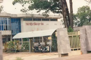 Bức ảnh chụp mặt tiền của một tòa nhà lớn, trên tường có dòng chữ màu đỏ 'THƯ VIỆN TỔNG HỢP TỈNH'. Đây là một công trình công cộng, có kiến trúc bê tông khá hiện đại vào thời điểm năm 1997, nổi bật với các ô cửa sổ kính lớn màu xanh. Phía trước tòa nhà là một khoảng sân, được bao quanh bởi hàng rào kim loại và cổng trụ đá. Dưới một mái che lợp tôn đơn giản, có ít nhất hai chiếc xe máy đang được đỗ. Cảnh vật xung quanh có nhiều cây xanh, bao gồm cả những cây thông cao lớn. Gần cổng có một tấm biển nhỏ màu vàng, nhưng chữ viết không thể đọc rõ, chỉ có thể nhận ra chữ 'CƠ QUAN' ở dòng trên. Không có hoạt động rõ rệt của con người trong ảnh, cho thấy một không gian yên tĩnh.。（日本語訳）。大きな建物の正面を写した写真で、壁には赤い文字で「THƯ VIỆN TỔNG HỢP TỈNH」（省総合図書館）と書かれています。これは公共の建物で、1997年当時としてはかなり近代的なコンクリート建築であり、大きな青いガラス窓が特徴的です。建物の前には庭があり、金属製の柵と石柱の門で囲まれています。シンプルなトタン屋根の庇の下に、少なくとも2台のバイクが停められています。周囲の景色には緑の木々が多く、背の高い松の木も含まれています。門の近くに小さな黄色い看板がありますが、文字ははっきりと読み取れず、上の行にある「CƠ QUAN」（機関）という文字だけが認識できます。写真には明確な人の活動は見られず、静かな空間であることがうかがえます。