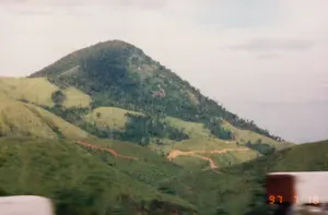 Dấu ngày tháng được ghi ở góc dưới bên phải là '97 7 18'.。Bức ảnh ghi lại phong cảnh một vùng đồi núi rộng lớn, có thể là khu vực Tây Nguyên hoặc trên tuyến đường từ Pleiku đến Nha Trang. Bầu trời u ám, có nhiều mây. Trọng tâm của bức ảnh là một ngọn núi hình nón, cùng với các ngọn đồi xung quanh được bao phủ bởi thảm cỏ xanh và các mảng rừng cây. Một con đường đất màu nâu đỏ uốn lượn quanh co trên sườn đồi. Ở phía xa, có thể nhận ra một cột điện cao thế. Không có người hay hoạt động cụ thể nào được nhìn thấy. Phần tiền cảnh bị mờ cho thấy bức ảnh có thể được chụp từ một phương tiện đang di chuyển.。（日本語訳）。右下に記録された日付は「97 7 18」です。写真は広大な丘陵地帯の風景を記録しており、おそらくテイグエン地方、またはプレイクからニャチャンへの道中のものです。空は曇っており、雲が多いです。写真の中心は円錐形の山で、その周りの丘は緑の草の絨毯と森の木々の斑点で覆われています。赤褐色の未舗装の道が丘の斜面を曲がりくねって続いています。遠くには、高圧電線の鉄塔が見えます。人や特定の活動は見られません。前景がぼやけていることから、写真が移動中の乗り物から撮影された可能性が示唆されます。