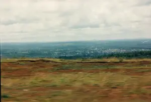 Bức ảnh chụp toàn cảnh một vùng thung lũng hoặc cao nguyên rộng lớn, nhìn từ trên cao. Tiền cảnh bị nhòe do chuyển động, cho thấy khả năng ảnh được chụp từ một phương tiện đang di chuyển. Phần đất ở tiền cảnh có màu nâu đỏ đặc trưng của đất bazan vùng Tây Nguyên, trên đó có mọc một ít cỏ xanh. Ở phía xa, một khu dân cư, có thể là một thị trấn hoặc một phần của thành phố, trải dài với nhiều mái nhà san sát. Bao quanh khu dân cư là những mảng rừng cây xanh và đất nông nghiệp. Phía sau nữa, ở đường chân trời, là những dãy núi nhấp nhô mờ ảo. Bầu trời u ám, đầy mây trắng và xám. Không có bất kỳ văn bản nào có thể đọc được trong ảnh.。（日本語訳）。高所から撮影された、広大な渓谷または高原地帯の全景写真です。前景は動きによってぼやけており、移動中の乗り物から撮影された可能性を示しています。前景の地面は、テイグエン地方の玄武岩土壌に特有の赤褐色をしており、その上には緑の草が少し生えています。遠方には、町または都市の一部と思われる住宅地が、多くの屋根を密集させながら広がっています。住宅地の周りには、緑の森林地帯と農地が広がっています。さらにその後方、地平線には、ぼんやりとした起伏のある山脈が見えます。空は曇っており、白と灰色の雲で覆われています。写真には読み取れる文字は一切ありません。