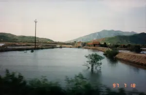 Bức ảnh được chụp từ bên trong một chiếc ô tô đang di chuyển trên một con đường nông thôn hoặc quốc lộ ở Việt Nam, với dấu ngày tháng '97 7 18' ở góc dưới bên phải. Ở phía trước, một chiếc xe tải màu trắng đang chạy ngược chiều, trên nóc xe chất đầy hàng hóa. Hai bên đường là lề đất, cây cối và bụi rậm mọc um tùm. Phía xa bên trái có thể thấy một vài ngôi nhà, và bên phải có các cột điện. Bầu trời u ám và nhiều mây. Có một vài người đang đứng hoặc đi bộ bên lề đường. Phần dưới của bức ảnh cho thấy cần gạt nước và một phần của bảng điều khiển của chiếc xe nơi người chụp đang ngồi.。（日本語訳）。写真はベトナムの田舎道または国道を走行中の車内から撮影されたもので、右下隅に「'97 7 18」の日付スタンプがあります。前方では、白いトラックが対向車線を走っており、その屋根には荷物が満載されています。道路の両側は土の路肩で、木々や茂みが鬱蒼と生い茂っています。遠くの左側には数軒の家が見え、右側には電柱があります。空はどんよりとしており、雲が多いです。路肩には数人が立っているか、歩いています。