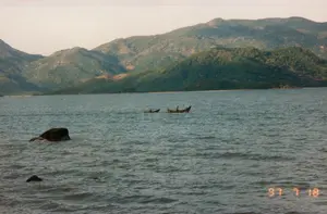 Dấu ngày tháng được in ở góc dưới bên phải bức ảnh là '97 7 18'. Bối cảnh là một vùng biển hoặc vịnh rộng lớn, có thể là ở khu vực Nha Trang. Phía xa là dãy núi trập trùng được bao phủ bởi cây xanh. Mặt nước biển gợn sóng. Trên mặt biển có hai chiếc ghe nhỏ, dường như là ghe gỗ truyền thống, đang di chuyển. Có thể thấy bóng người trên một trong hai chiếc ghe, có khả năng là ngư dân đang hoạt động đánh bắt hoặc di chuyển trên biển. Ở phía tiền cảnh có những tảng đá nhô lên khỏi mặt nước.。（日本語訳）。写真の右下に印刷された日付は「'97 7 18」です。背景は広大な海または湾で、おそらくニャチャン地域でしょう。遠方には緑に覆われた起伏のある山脈があります。海面は波立っています。海上には、伝統的な木造船と思われる2隻の小さな船が移動しています。2隻の船のうちの1隻に人影が見え、漁師が漁をしているか、海上を移動している可能性があります。前景には水面から突き出た岩があります。