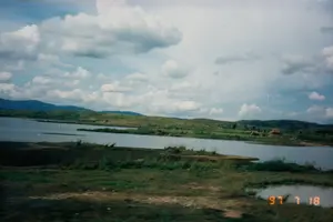 Khung cảnh chụp một vùng vịnh ven biển, có thể là một làng chài gần Nha Trang. Trên mặt biển yên tĩnh, có rất nhiều thuyền đánh cá đang neo đậu. Các loại thuyền bao gồm cả ghe gỗ nhỏ truyền thống và ít nhất một chiếc tàu lớn hơn có cabin, trên tàu có cắm một lá cờ đỏ. Một số thuyền khác được kéo lên bãi cát. Dọc bờ biển là một vài ngôi nhà đơn sơ nằm giữa cây cối, dưới chân một ngọn đồi xanh. Phía xa là những dãy núi trập trùng. Ở tiền cảnh, có một bức tường kè xây bằng đá và một khoảng sân đất trống có trồng vài cây non. Một người phụ nữ đang đứng gần bờ biển. Góc dưới cùng bên phải của bức ảnh có ghi ngày tháng '97 7 18', cho thấy thời điểm chụp là ngày 18 tháng 7 năm 1997.。（日本語訳）。沿岸の湾を撮影した風景で、おそらくニャチャン近郊の漁村だろう。静かな海面には、たくさんの漁船が停泊している。他のいくつかの船は砂浜に引き上げられている。海岸沿いには、緑の丘の麓、木々の間にいくつかの質素な家が建っている。遠くには、起伏のある山々が連なっている。前景には、石で造られた擁壁と、いくつかの若木が植えられた空き地がある。一人の女性が海岸の近くに立っている。写真の右下隅には「'97 7 18」という日付が記されており、撮影日時が1997年7月18日であることを示している。