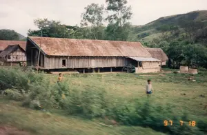 Bức ảnh chụp một chiếc xe tải màu trắng có mui bạt màu xanh đậm đang chạy trên đường, ngang qua một trạm kiểm tra. Trên mái ngói của tòa nhà có cắm một lá cờ Việt Nam (cờ đỏ sao vàng). Có một tấm biển hiệu màu đỏ chữ vàng trên tòa nhà, ghi dòng chữ "TRẠM KIỂM TRA" ở trên và "XE QUA..." ở dưới, phần còn lại của dòng chữ bị cabin xe tải che khuất nên không thể đọc được. Có một người đang ngồi trong cabin xe tải. Phía sau trạm kiểm tra là khung cảnh đồi núi.。（日本語訳）。濃い青色の幌が付いた白いトラックが、検問所を通り過ぎて道路を走っている写真です。建物の瓦屋根の上には、ベトナムの国旗（金星紅旗）が掲げられています。建物には赤地に黄色の文字で書かれた看板があり、上段に「TRẠM KIỂM TRA」、下段に「XE QUA...」と書かれていますが、残りの文字はトラックの運転台に隠れて読むことができません。トラックの運転台には人が一人座っています。検問所の後ろには、丘や山の風景が広がっています。