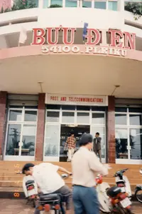 Bức ảnh chụp mặt tiền của Bưu điện Pleiku vào năm 1997. Trên mái hiên cong của tòa nhà có một biển hiệu lớn với dòng chữ "BƯU ĐIỆN" màu đỏ, bên dưới là "54100 . PLEIKU" màu trắng. Ngay phía trên cửa ra vào chính có một biển hiệu khác bằng tiếng Anh ghi "POST AND TELECOMMUNICATIONS". Đây là một tòa nhà công cộng, có kiến trúc hiện đại với nhiều tầng và cửa kính. Phía trước tòa nhà là các bậc thang dẫn lên lối vào. Có một vài người đang hoạt động: một người đàn ông mặc áo sơ mi kẻ sọc đang đi ra từ cửa, một người đàn ông khác mặc vest đang đứng gần đó. Ở tiền cảnh, một người đàn ông mặc áo phông trắng đang ngồi trên xe máy. Một số xe máy kiểu dáng phổ biến của thập niên 90 đang được dựng ở phía trước. Khung cảnh thể hiện hoạt động thường ngày tại một bưu điện ở thành phố Việt Nam.。（日本語訳）。1997年に撮影されたプレイク郵便局の正面の写真です。建物の湾曲した庇の上には、赤い文字で「BƯU ĐIỆN」と書かれた大きな看板があり、その下には白い文字で「54100 . PLEIKU」と書かれています。正面玄関のすぐ上には、英語で「POST AND TELECOMMUNICATIONS」と書かれた別の看板があります。これは公共の建物で、複数の階とガラスのドアを持つ現代的な建築様式です。建物の前には、入り口へと続く階段があります。数人の人々が活動しています。縞模様のシャツを着た男性がドアから出てきており、別のベストを着た男性がその近くに立っています。前景には、白いTシャツを着た男性がバイクに座っています。90年代に一般的だったデザインのバイクが数台、前に停められています。この光景は、ベトナムの都市にある郵便局での日常の活動を表しています。