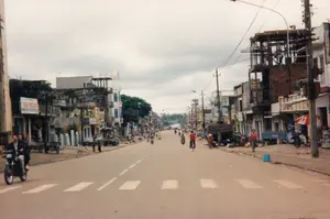 Đây là quang cảnh một con đường phố tại một thị trấn ở Việt Nam vào năm 1997. Con đường được trải nhựa và có vạch kẻ cho người đi bộ. Hai bên đường là những dãy nhà phố, cửa hàng san sát nhau. Ở phía bên trái, có một cửa hàng với biển hiệu lớn màu trắng ghi chữ 'KIM THÀNH'. Có nhiều biển hiệu khác trên các tòa nhà nhưng không thể đọc rõ. Phía bên phải, một tòa nhà đang được xây dựng dang dở với khung giàn giáo và tường gạch. Hoạt động trên đường phố khá nhộn nhịp với nhiều người dân đi lại bằng xe máy và xe đạp. Có hai người đàn ông đang đi xe máy ở gần phía trước. Trên vỉa hè, đặc biệt là bên phải, có cảnh buôn bán nhỏ lẻ, một số người tụ tập quanh hàng hóa bày trên mặt đất, và một người phụ nữ đội nón lá. Các vật thể đáng chú ý khác bao gồm cột điện với dây điện chằng chịt, một chiếc xe tải nhỏ, một chiếc xe ba gác, và những chiếc xe kéo tay.。（日本語訳）。これは1997年、ベトナムのある町の通りの光景です。道路はアスファルトで舗装されており、横断歩道があります。道路の両側には、店舗が密集した家々が並んでいます。左側には、「KIM THÀNH」と書かれた大きな白い看板のある店があります。右側では、足場のフレームとレンガの壁がある建物が建設途中です。通りは、バイクや自転車で行き交う多くの人々でかなり賑わっています。手前近くには、バイクに乗っている2人の男性がいます。歩道、特に右側では、小規模な商売の光景が見られ、地面に並べられた商品の周りに数人が集まっており、ノンラーをかぶった女性が一人います。その他の特筆すべき物には、電線が絡み合った電柱、小型トラック1台、三輪車1台、そして数台の手押し車が含まれます。