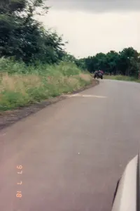 Trên ảnh có dấu ngày tháng "97 7 18".。Bối cảnh là một con đường nhựa ở vùng nông thôn. Con đường uốn cong nhẹ về phía xa. Ven đường là lề đất với cỏ dại mọc um tùm, và xa hơn là những hàng cây xanh rậm rạp. Bầu trời có nhiều mây.。Ở phía xa trên đường, có một chiếc xe trông giống xe công nông hoặc một loại xe cơ giới nhỏ tự chế, đang di chuyển cùng chiều. Có thể thấy vài người đang ngồi trên chiếc xe này. Bức ảnh dường như được chụp từ một phương tiện khác (có thể là ô tô) đang đi phía sau, với một phần thân xe màu sáng lọt vào khung hình ở góc dưới bên phải.。（日本語訳）。写真には「97 7 18」という日付スタンプがある。背景は田舎のアスファルト道路である。道は遠くに向かって緩やかにカーブしている。道端は雑草が生い茂る土の路肩で、その向こうには緑豊かな木々が鬱蒼と茂っている。空は雲が多い。道路の遠くには、耕運機か自家製の小型車両のような車が1台、同じ方向に進んでいる。この写真は、後方を走る別の乗り物（おそらく自動車）から撮影されたようで、右下の隅に明るい色の車体の一部がフレームに入り込んでいる。