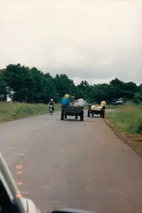 Trong ảnh có ghi ngày tháng là '97 7 18', tức là ngày 18 tháng 7 năm 1997.。Bối cảnh là một con đường nhựa ở vùng nông thôn, có thể là ở khu vực Tây Nguyên. Hai bên đường là cỏ và phía sau là những hàng cây xanh rậm rạp. Bầu trời có vẻ u ám, nhiều mây.。Hoạt động chính là cảnh sinh hoạt và giao thông trên đường. Có hai chiếc xe ngựa (hoặc xe bò) thô sơ đang di chuyển trên đường. Đây là phương tiện vận chuyển người và hàng hóa phổ biến ở nông thôn Việt Nam vào thời điểm đó.。- Chiếc xe ngựa đi trước chở khoảng ba người, trong đó có một người mặc áo xanh lam và đội nón lá.。- Chiếc xe ngựa thứ hai đi sau, chở đầy các bao tải màu trắng, có một người mặc áo vàng đi bên cạnh.。Ngoài ra, có một người đang lái xe máy đi ngược chiều với hai chiếc xe ngựa. Người lái xe máy đội mũ bảo hiểm. Bức ảnh dường như được chụp từ bên trong một chiếc ô tô đang di chuyển, vì có một phần của khung xe ở góc dưới bên trái.。（日本語訳）。写真には「'97 7 18」という日付が書かれています。つまり、1997年7月18日です。背景は田舎のアスファルト道路で、おそらくタイグエン地方でしょう。道の両側は草で、後ろには緑の木々が鬱蒼と茂っています。空はどんよりしているようで、雲が多いです。主な活動は、道路上の生活と交通の風景です。2台の簡素な馬車（または牛車）が道路上を移動しています。これは当時、ベトナムの田舎で人と荷物を運ぶ一般的な輸送手段でした。- 前を行く馬車は約3人を乗せており、その中には青いシャツを着てノンラー（菅笠）をかぶっている人がいます。- 後ろを行く2台目の馬車は、白い袋をたくさん積んでおり、黄色いシャツを着た人が横を歩いています。その他に、2台の馬車とは反対方向に走っているバイクを運転している人が一人います。バイクの運転手はヘルメットをかぶっています。この写真は、左下の隅に車体の一部が写っていることから、移動中の車内から撮影されたようです。