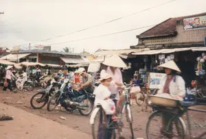 看板で識別できる文字は以下の通りです：。- スタンド看板：「NHẬN PHOTOCOPY」（コピー承ります）。- ある建物にある赤と黄色の大きな看板：「... BÁN SỈ LẺ ...」（...卸売・小売...）。- 遠くの家の屋根にある青い看板：「HỘI ĐỒNG ... NGHĨA TRINH」、一部の文字ははっきりと読み取れません。- 他のいくつかの看板はぼやけており、読み取ることができません。これはベトナムの町または農村部にある屋外市場の光景です。主要な道は土の道で、ぬかるんでいるように見えます。主な活動は、人々の売買や賑やかな往来の光景です。多くの人々がバイク（主に古いモデル）や自転車で移動しています。手前では、ノンラーをかぶった女性が、自転車の前に取り付けられた小さな椅子に座っている子供を乗せています。その隣では、同じくノンラーをかぶった別の女性が自転車を押しており、後ろには露天販売用の商品を乗せた大きな盆があります。二人の男性がバイクに座っています。市場の中では、他の多くの人々が歩いたり、仮設の屋根がある露店で買い物をしたりしています。目立つ物としては、多くのノンラー、その時代に普及していた種類のバイクや自転車、そして瓶詰めの清涼飲料水やおもちゃなどの多くの商品を売る簡素な露店などがあります