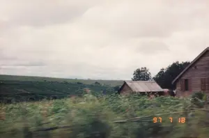 Trong ảnh có ghi ngày tháng '97 7 18'. Bức ảnh chụp một khung cảnh nông thôn ở vùng cao nguyên, có thể là trên đường đi giữa Pleiku và Nha Trang. Cảnh vật cho thấy hai ngôi nhà đơn sơ. Một ngôi nhà lớn hơn ở bên phải được làm bằng gỗ, có cửa sổ chớp bằng gỗ. Ngôi nhà nhỏ hơn ở bên trái có mái lợp tôn. Phía sau những ngôi nhà là một vùng đồi núi rộng lớn, được bao phủ bởi các cánh đồng hoặc đồn điền cây công nghiệp xanh mướt, có khả năng là cây cà phê hoặc chè, đặc trưng của vùng Tây Nguyên. Phần tiền cảnh của bức ảnh bị nhòe do chuyển động, cho thấy nó được chụp từ một phương tiện đang di chuyển. Bầu trời u ám và đầy mây.。（日本語訳）。写真には「'97 7 18」という日付が記されている。この写真は高原地帯の田舎の風景を撮影したもので、おそらくプレイクとニャチャンの間の道中であろう。風景には2軒の質素な家が写っている。右側のより大きな家は木造で、木製の鎧戸窓がある。左側のより小さな家はトタン屋根である。家々の後ろには広大な丘陵地帯が広がり、緑豊かな畑や換金作物のプランテーションで覆われている。それはおそらく、西ハイランド地方特有のコーヒーの木か茶の木であろう。写真の前景は動きによってぼやけており、移動中の乗り物から撮影されたことを示している。空はどんよりとしており、雲に覆われている。