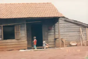 この写真は、農村地域にある一軒の木造家屋の前での生活風景を記録したもので、右下の「\'97 7 18」という日付表示から1997年7月18日に撮影されたことがわかります。場所は特徴的な赤土の地面で、テイグエン地方の田舎である可能性があります。家はシンプルな建築様式で、壁は木の板で、屋根は素焼きの瓦で葺かれています。家の前では二人の子供が遊んでいます。赤い服を着た一人の子供が戸口に立っています。もう一人の子供は帽子をかぶり、地面にしゃがんでいます。家の壁には、ほうき、数本の竹竿、竹で編んだ大きな平たいざるなどの農具が立てかけられており、ここが農家の家である可能性を示しています
